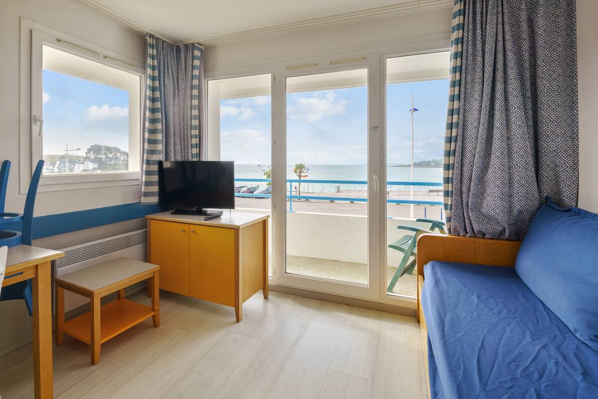 Ein Zimmer mit einem Holztisch, einem Fernseher, einer Couch und Meerblick durch große Glastüren.