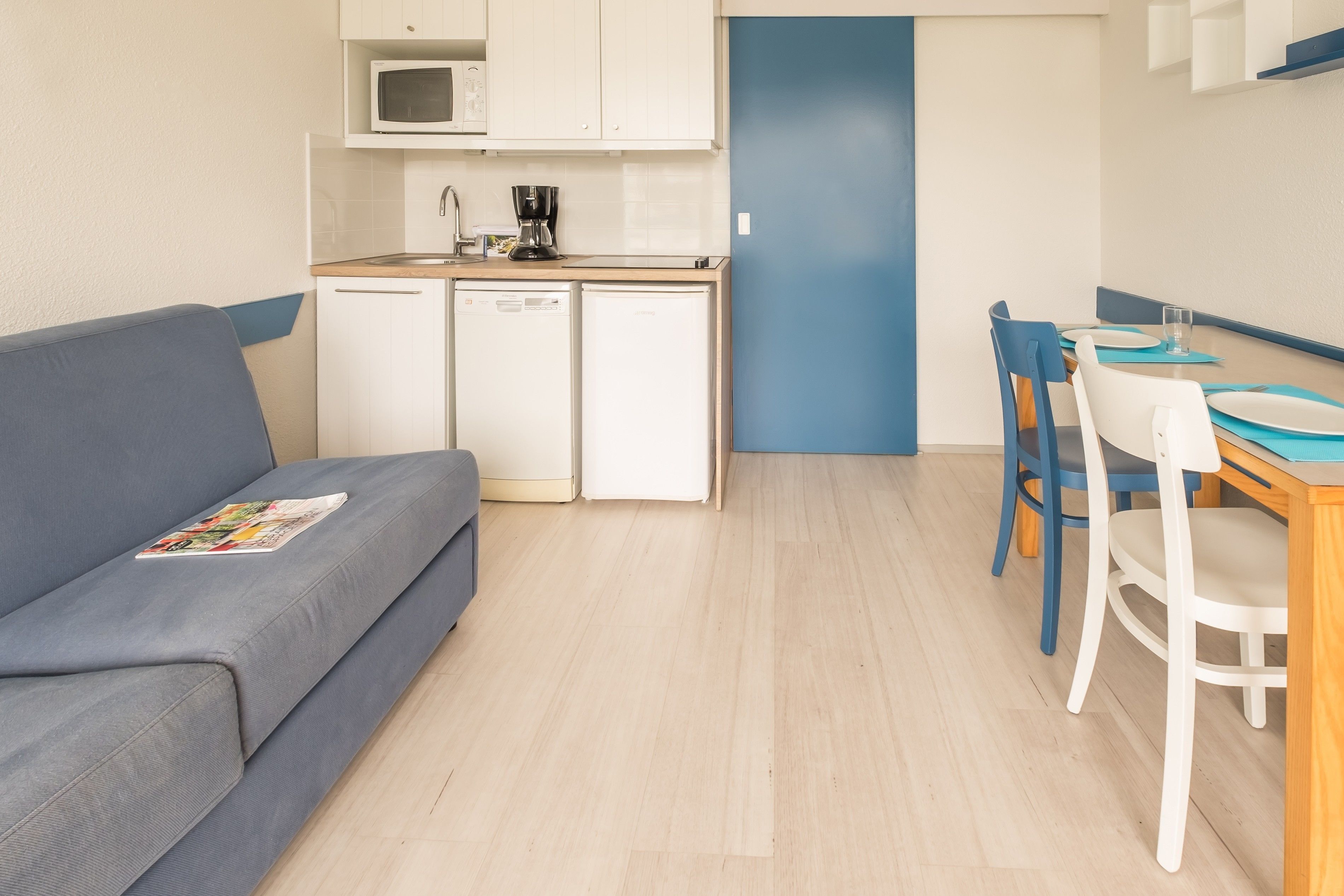 Um espaço compacto com cozinha compacta, área de refeições com cadeiras azuis e um sofá cinzento.