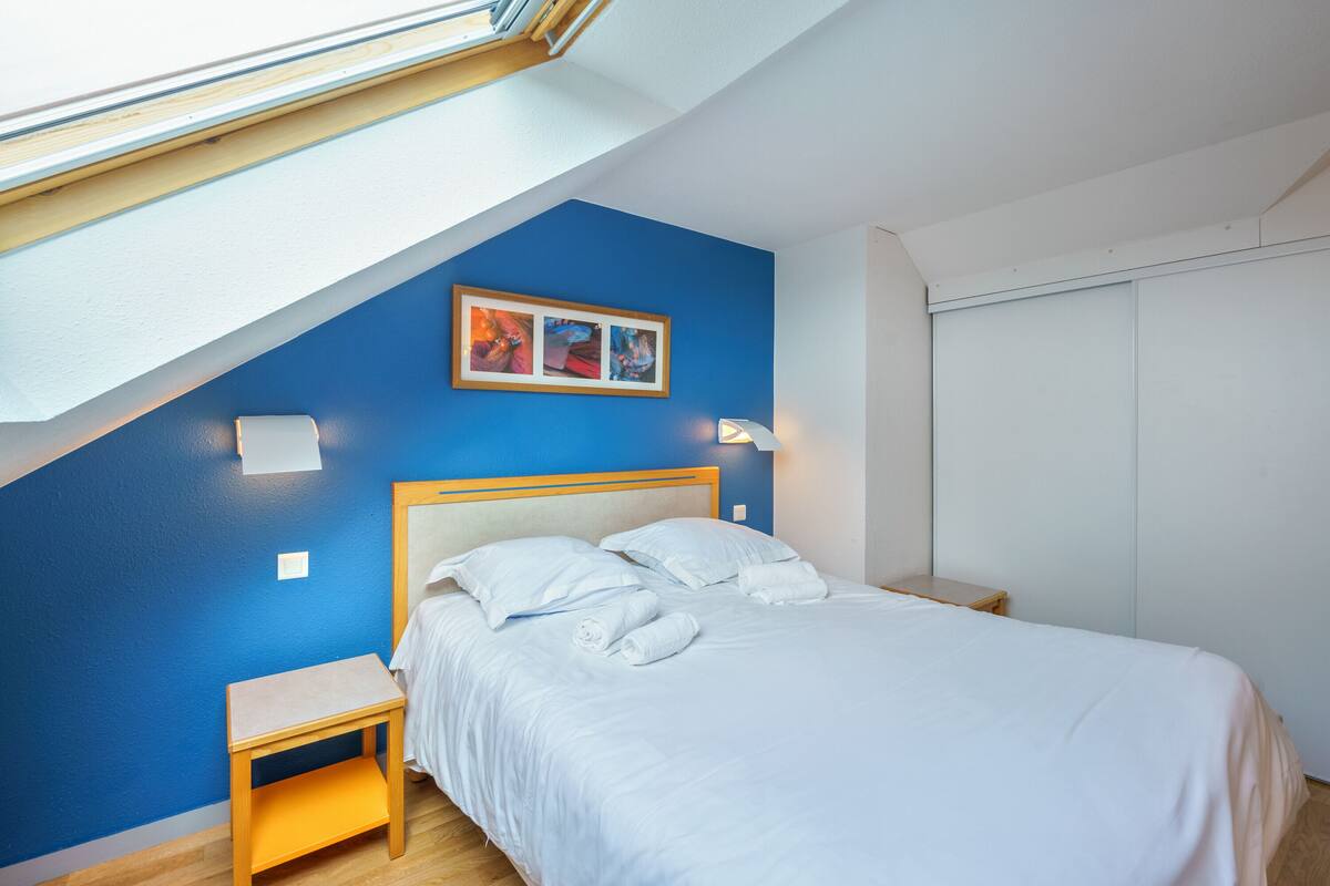 Ein Schlafzimmer mit Dachfenster, einem Bett mit weißen Leinen, einem Nachttisch und einer wandmontierten Lampe.