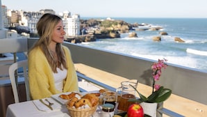 A table with breakfast items, a potted plant, and a view of the ocean and buildings. - Résidence Victoria Surf (Biarritz)