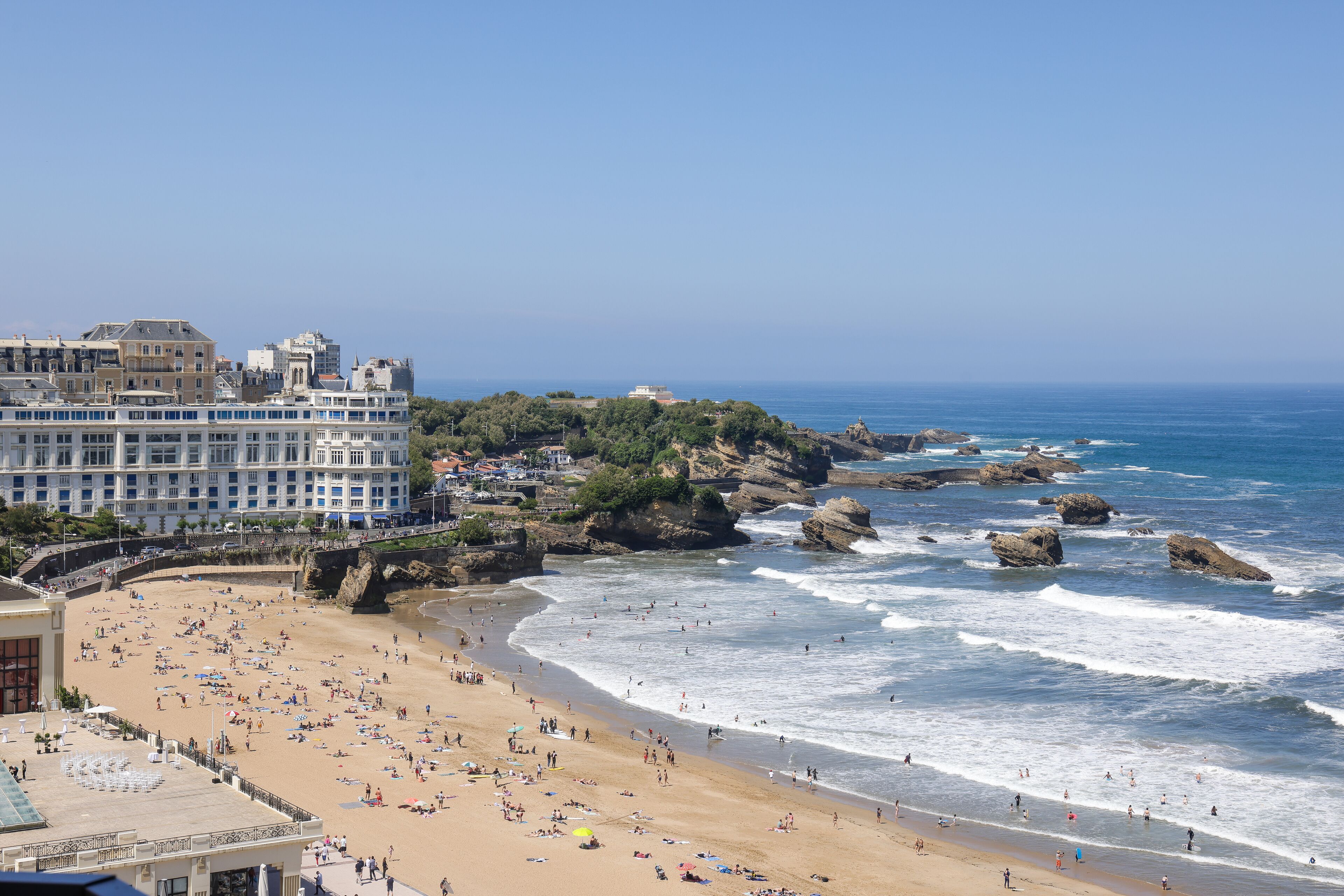 A beach with a hotel in the background, rocky coastline, and clear blue sky.