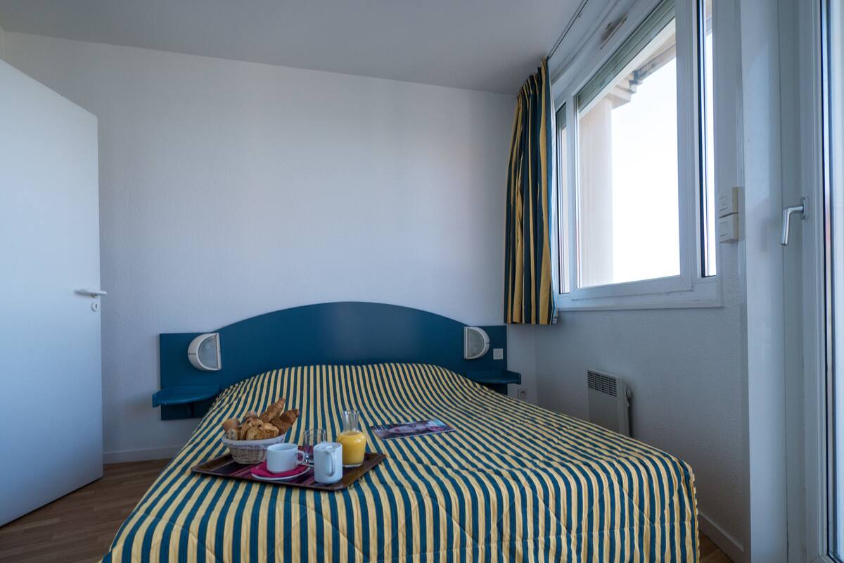 A bedroom with a bed, a window with curtains, a bedside table, and a wall-mounted clock.