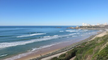 A coastal beach with waves, a rocky shoreline, and buildings in the background.