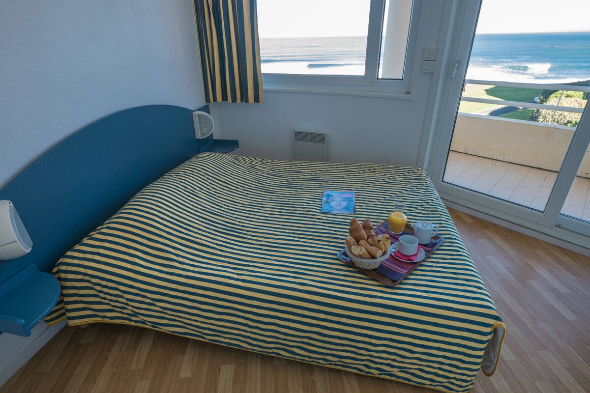 A room with a bed, a window with a view of the ocean, a bedside table with a lamp, and a tray with breakfast items.
