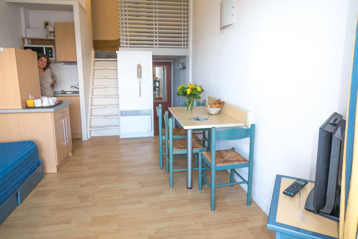 A compact living space with a kitchenette, a dining area with a table and chairs, a small TV, and a bed.