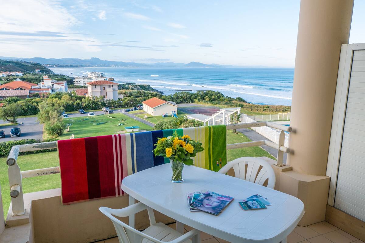 A balcony with a white table, chairs, and a view of the ocean and distant buildings.