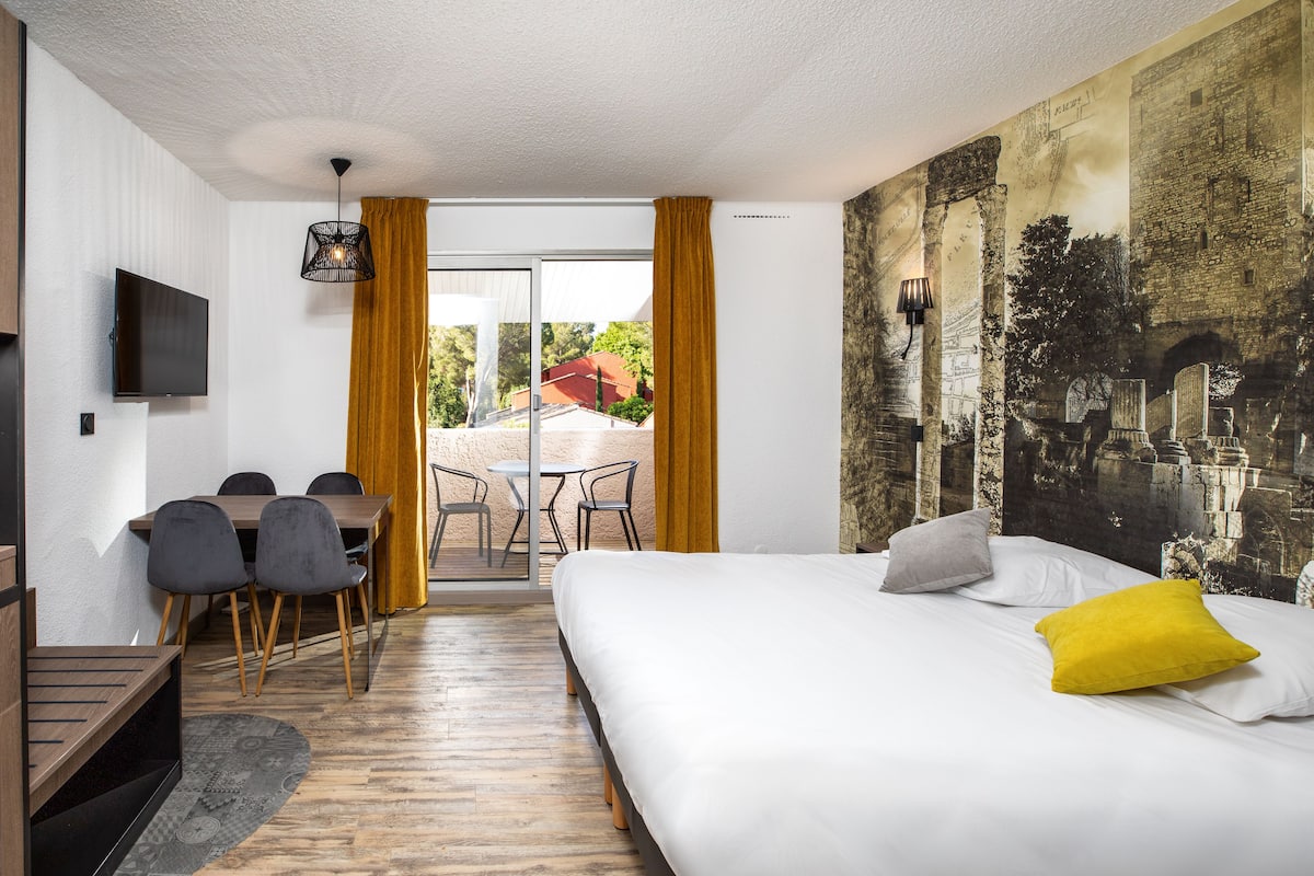 A hotel room with a bed, a desk with chairs, a TV, and a mural on the wall.