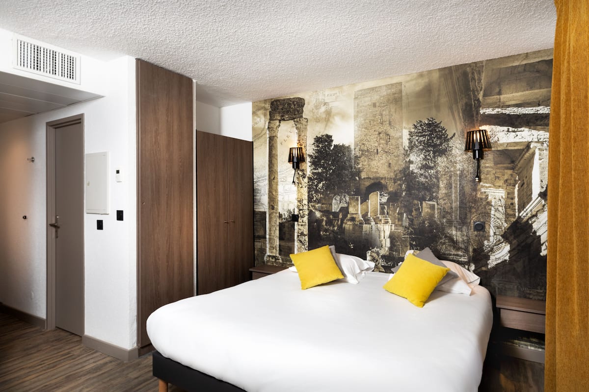 A hotel room with a large bed, two yellow pillows, a mural of ancient architecture on the wall, and a wooden wardrobe.