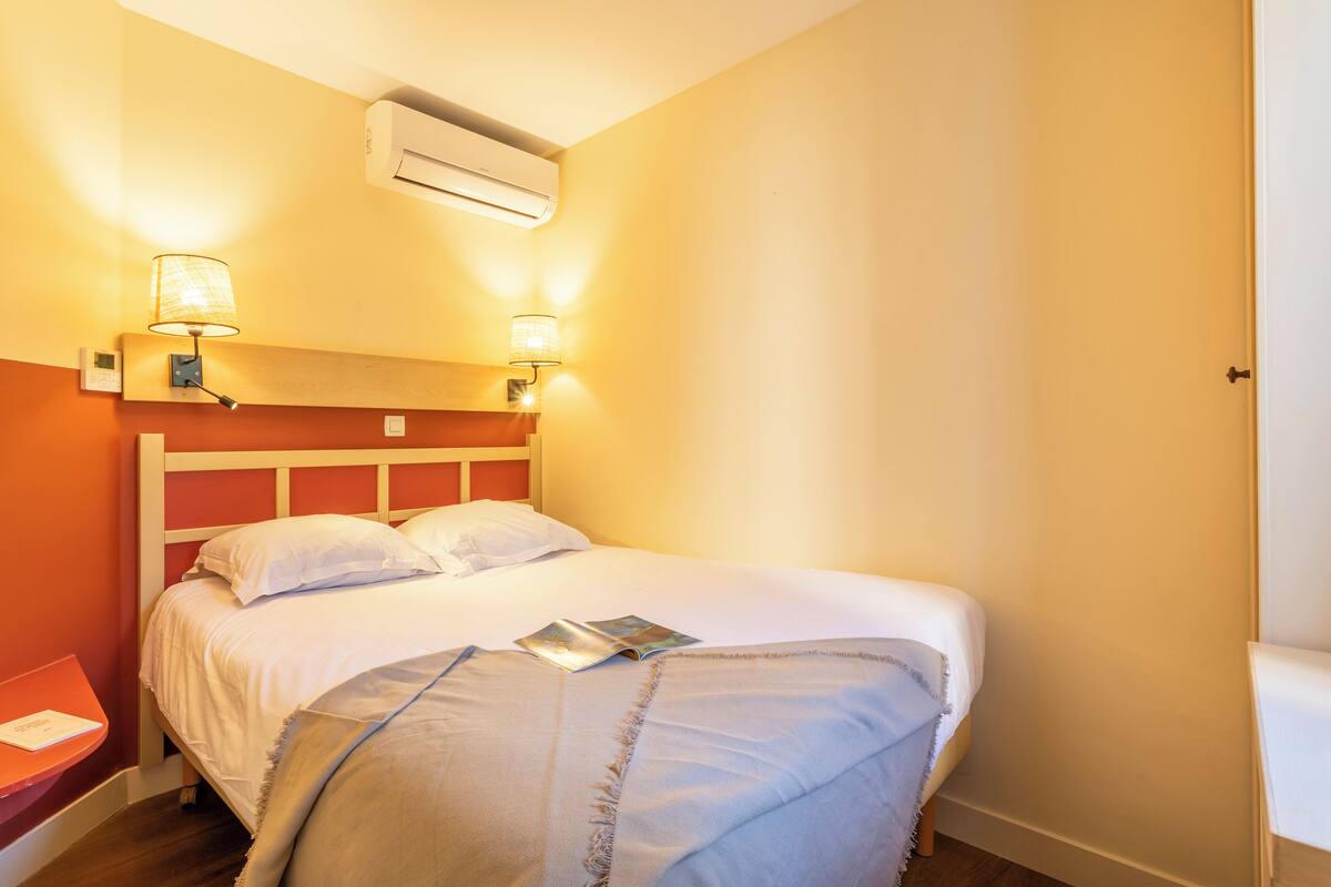 Un lit bien fait, avec du linge de lit blanc et un magazine posé dessus, une table de chevet avec une lampe et un appareil de climatisation au-dessus du lit.