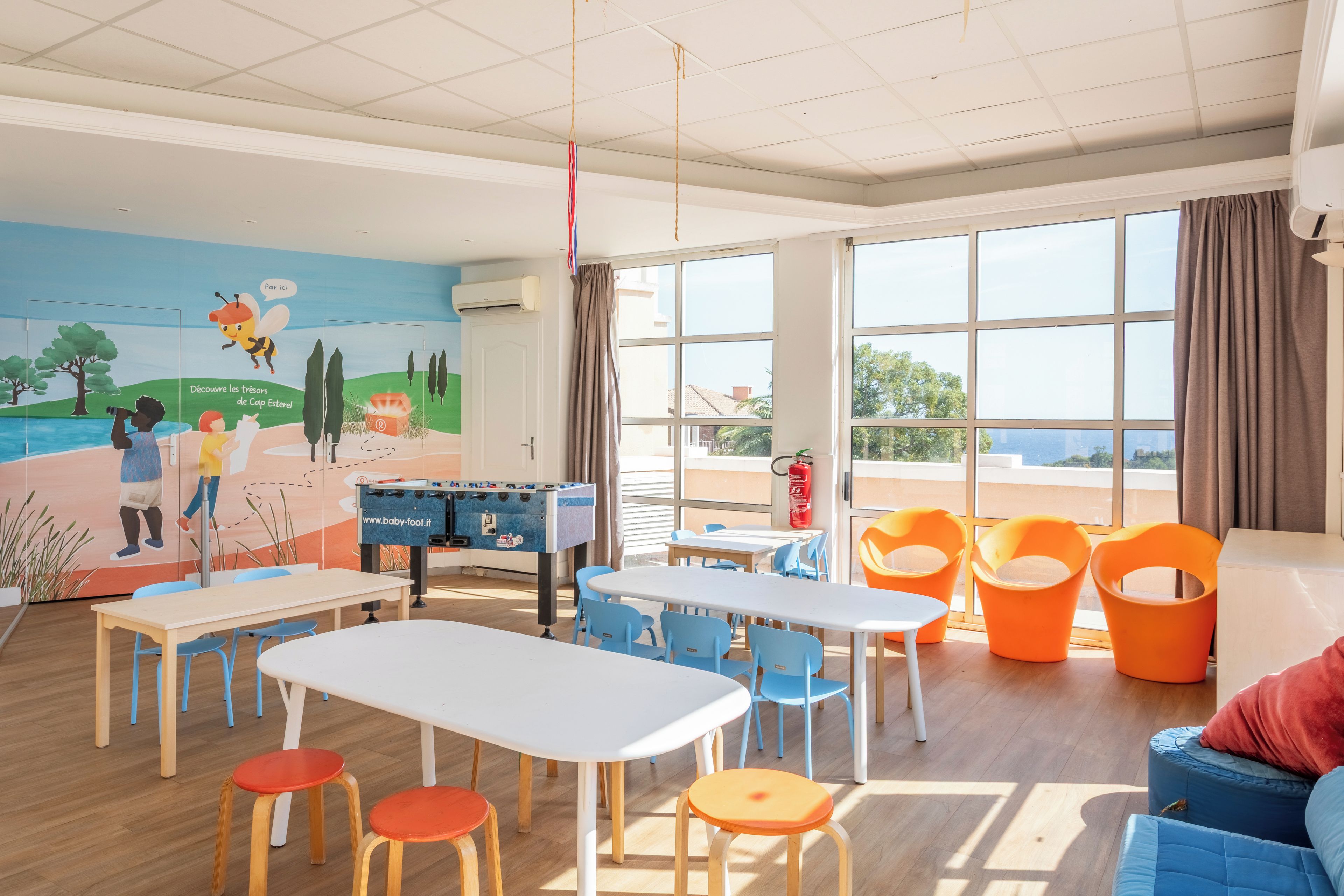 Una sala de juegos infantil con mesas, sillas, una mesa de fútbol de mesa y un mural en la pared.