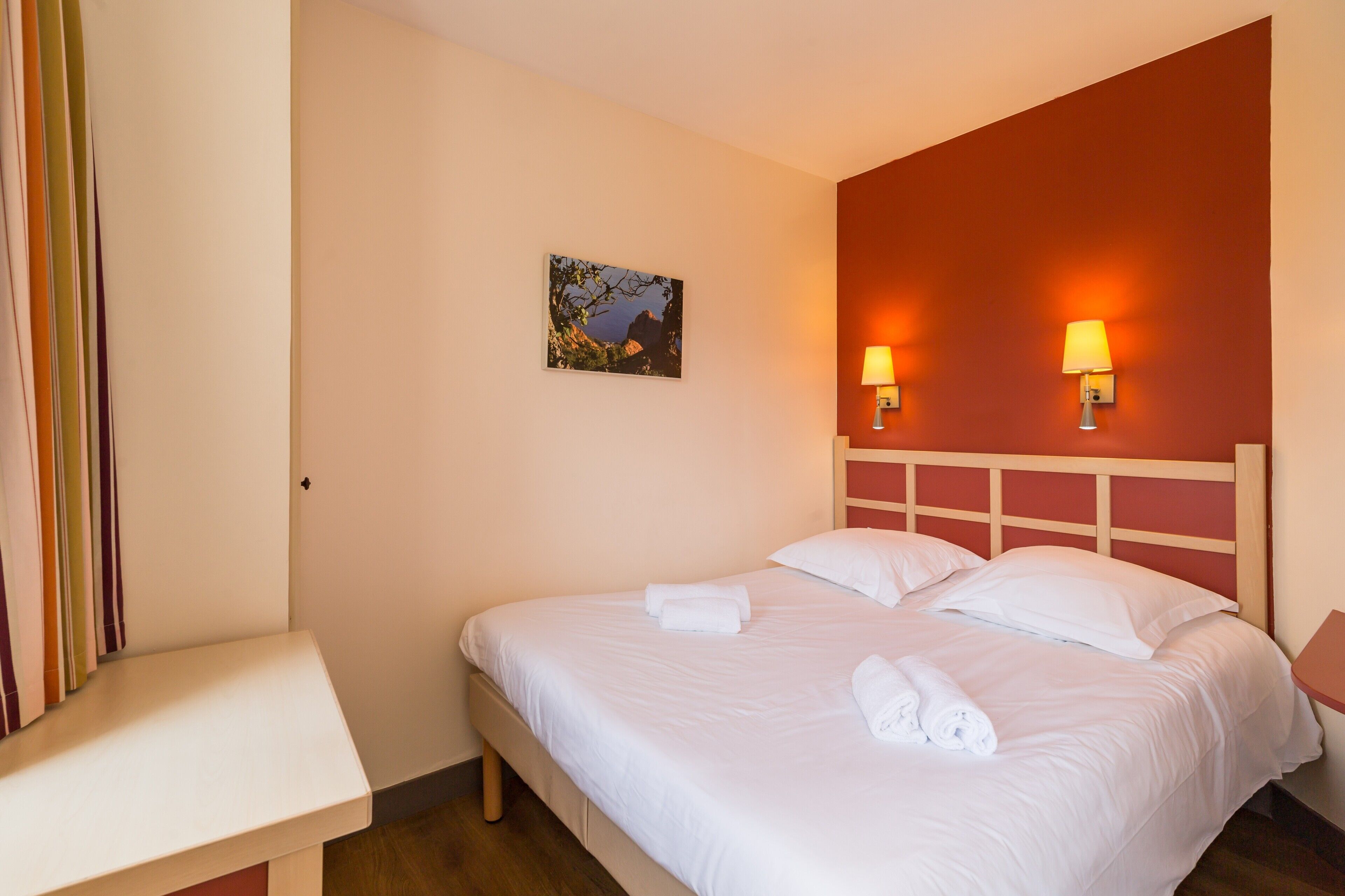 A neatly made bed with white linens and towels, a small bedside table, and two wall-mounted lamps.
