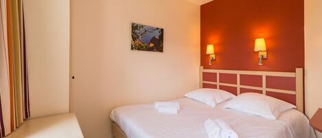 A neatly made bed with white linens and towels, a small bedside table, and two wall-mounted lamps.