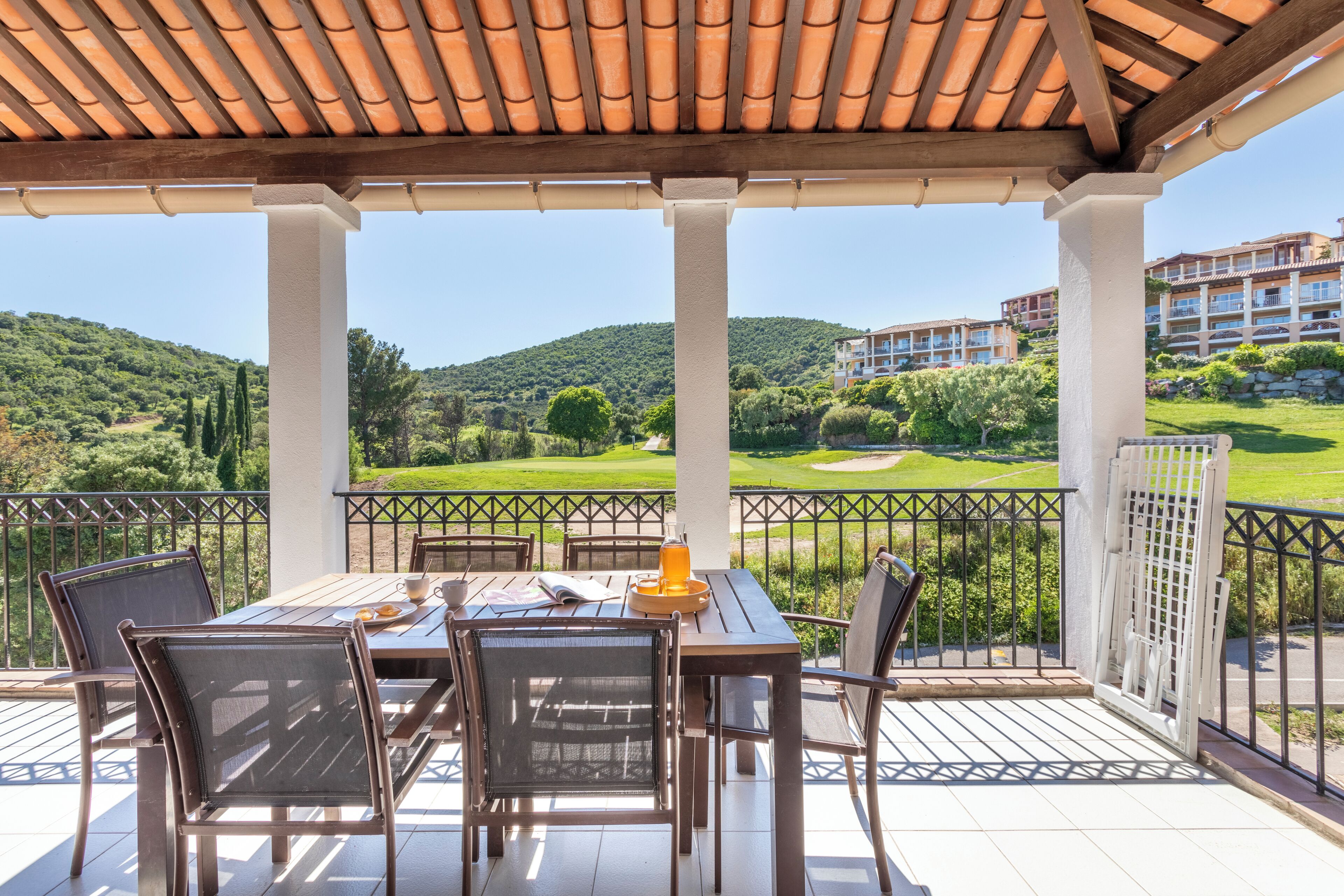 A covered outdoor seating area with a table and chairs, overlooking a golf course and residential buildings.