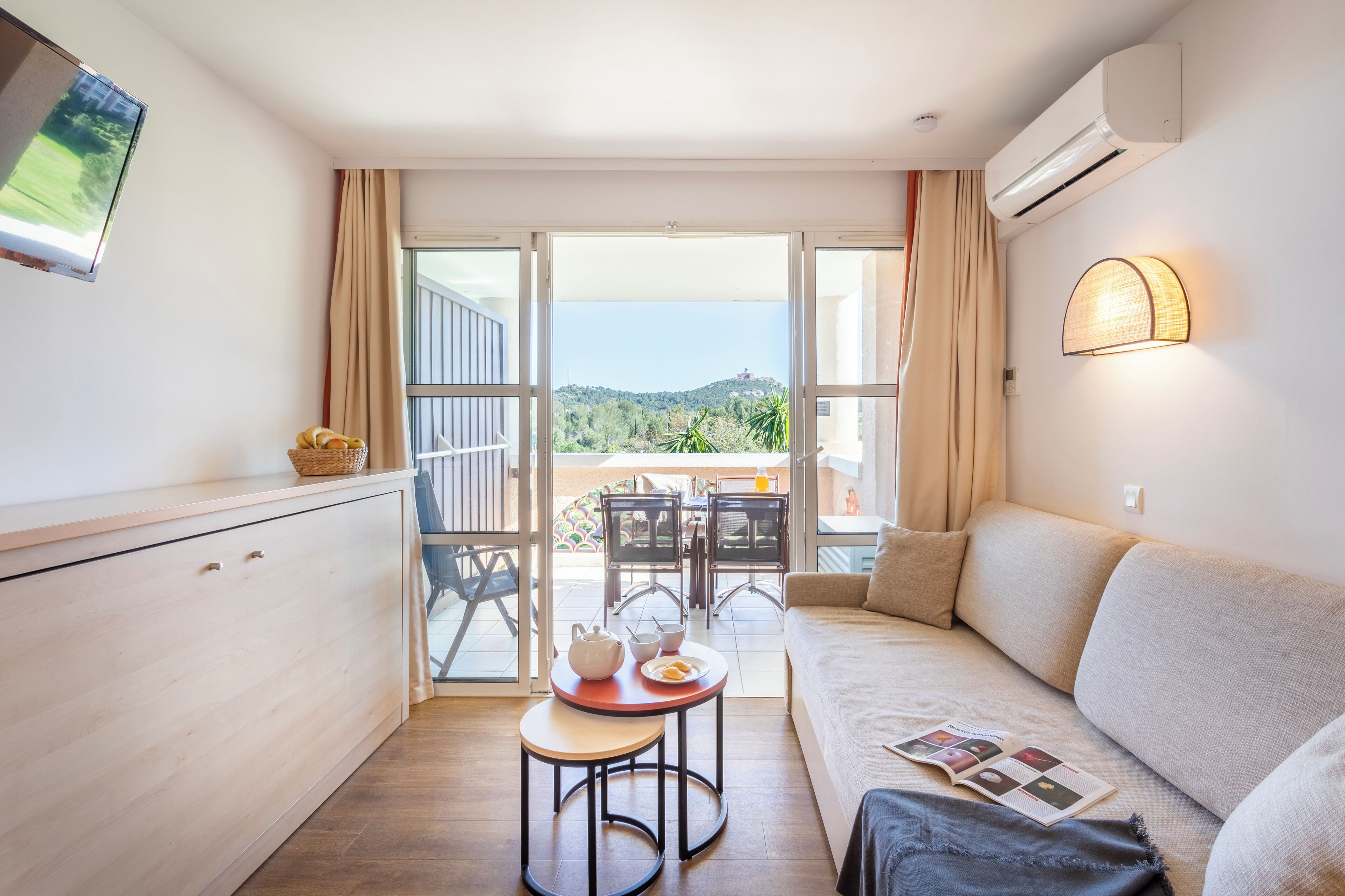 A modern room with a sofa, a small dining table with a teapot and cups, a TV mounted on the wall, and a balcony with a view of greenery and hills.