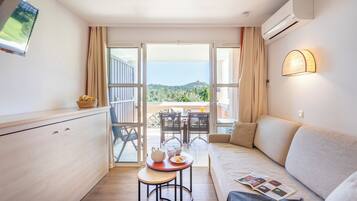 A modern room with a sofa, a small dining table with a teapot and cups, a TV mounted on the wall, and a balcony with a view of greenery and hills.