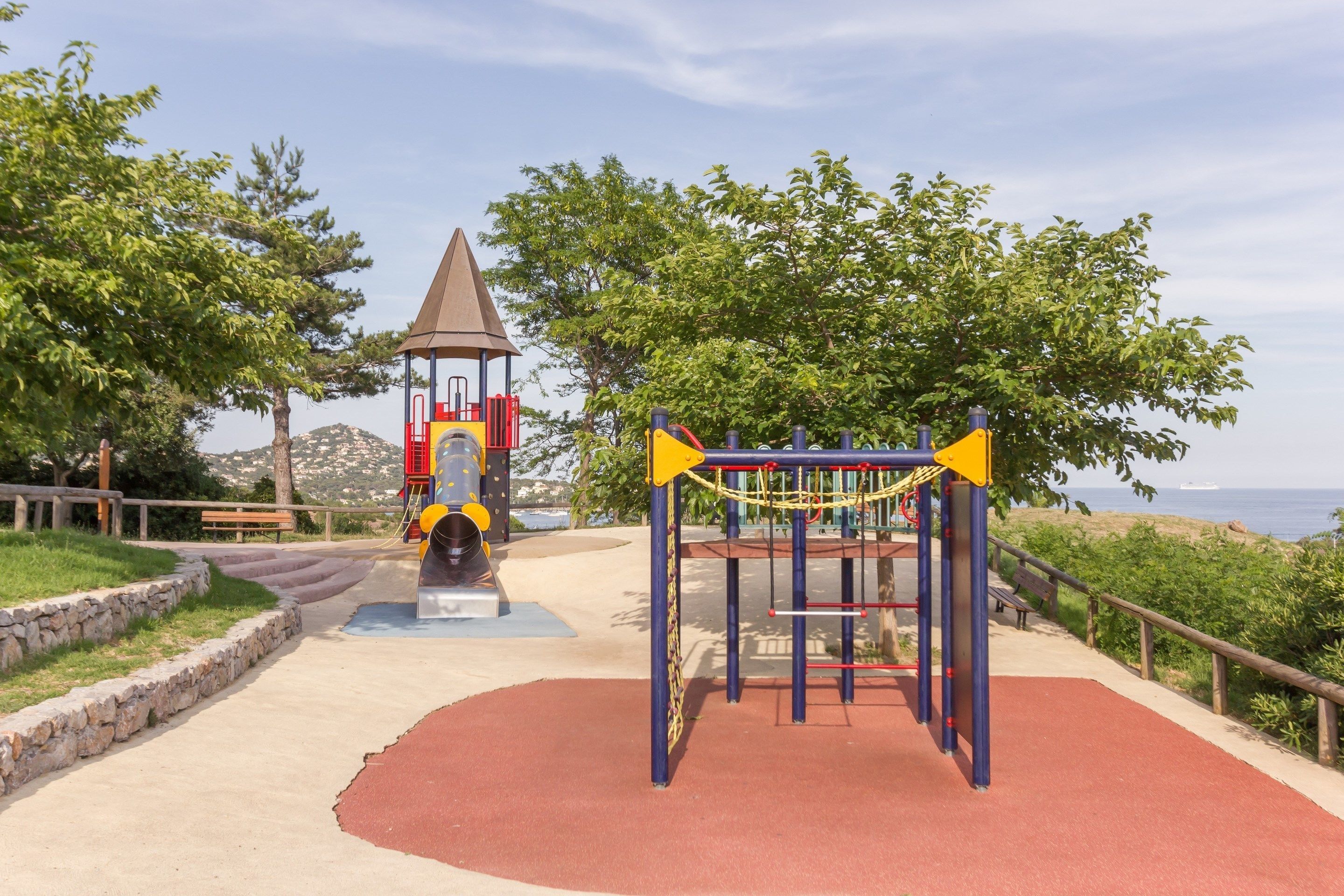 Un parque infantil con estructuras para escalar y un tobogán, rodeado de árboles y vistas al mar.