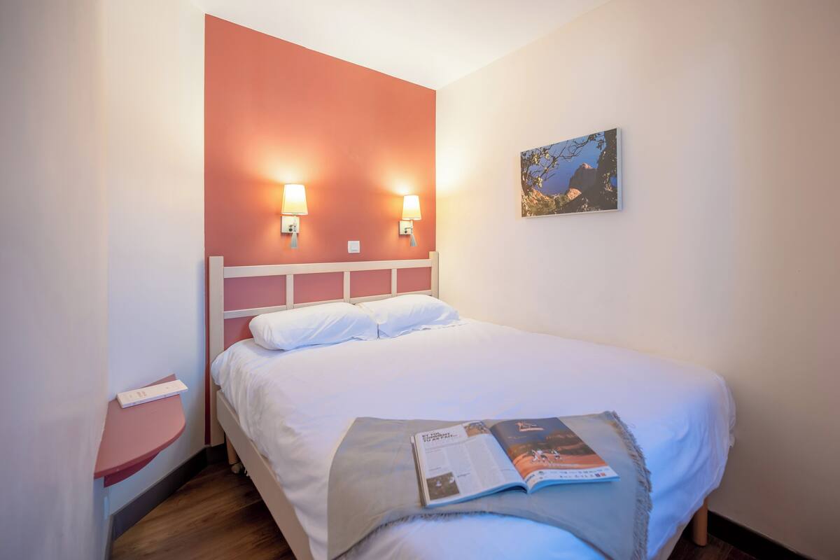 Un lit bien fait, avec une literie blanche, deux lampes fixées au mur et un tableau encadré accroché au mur.