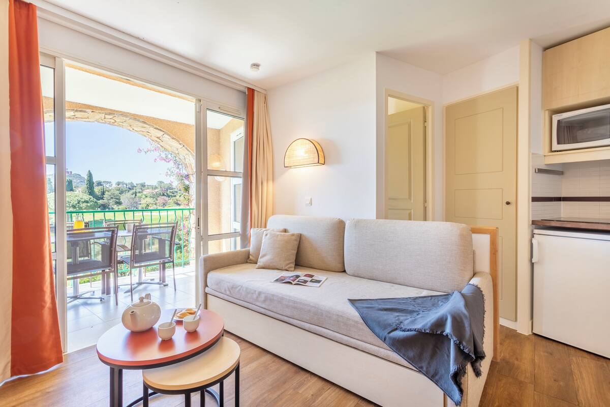 Un salon moderne avec un canapé, une petite table sur laquelle se trouvent une théière et des tasses, et une vue sur un balcon aménagé avec du mobilier d’extérieur.