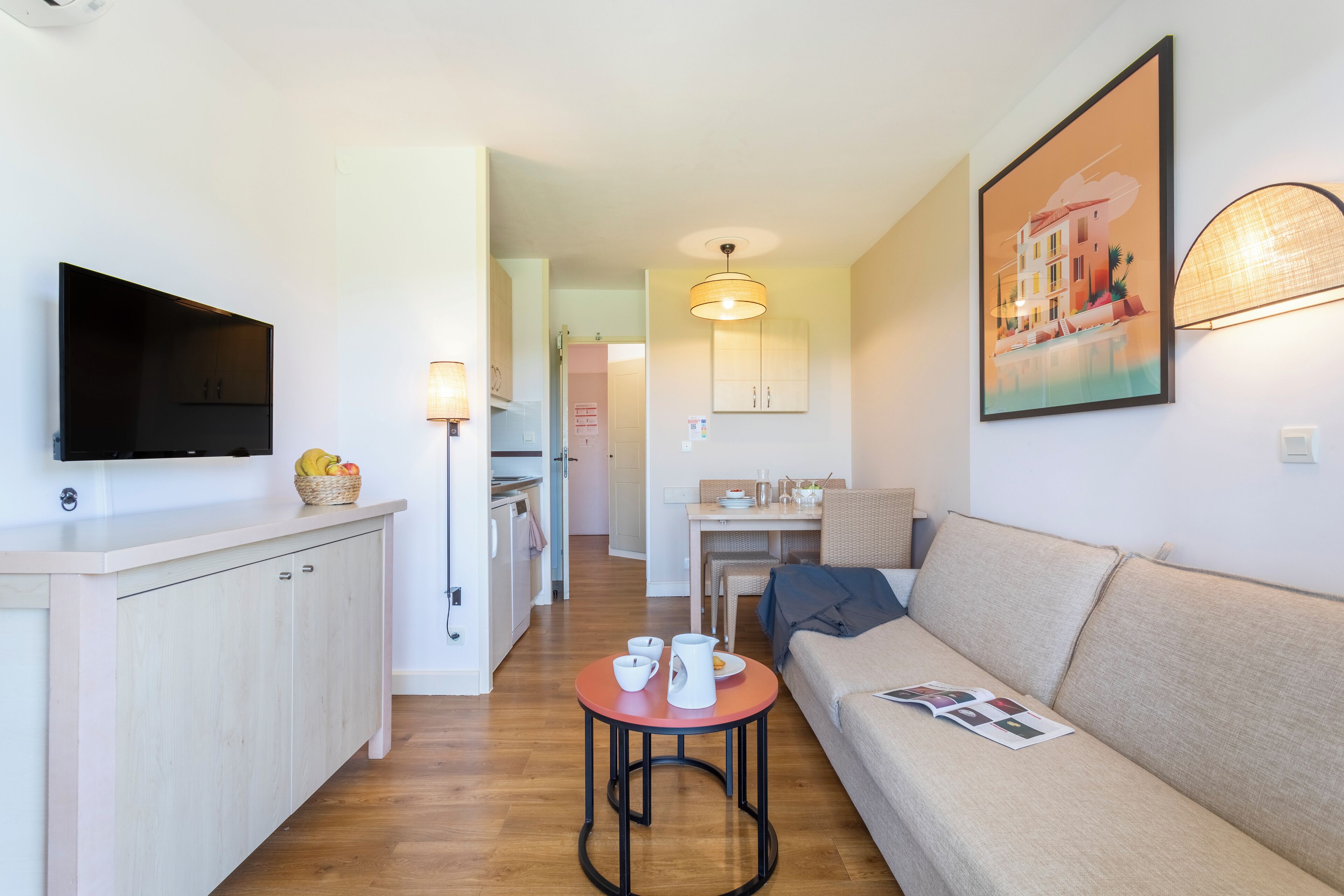 A modern living room with a sofa, a TV mounted on the wall, a dining table, and a kitchen area in the background.