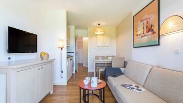 A modern living room with a sofa, a TV mounted on the wall, a dining table, and a kitchen area in the background.