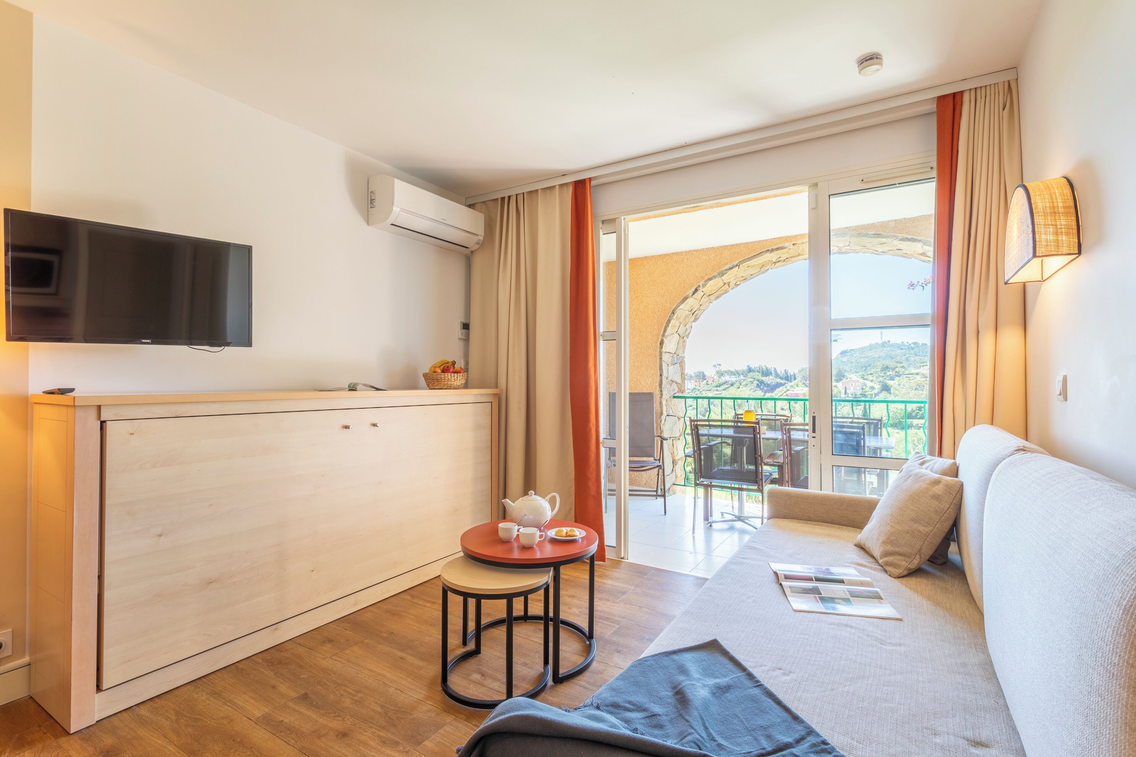 A modern room with a kitchen area, a sofa, and a balcony with a view.