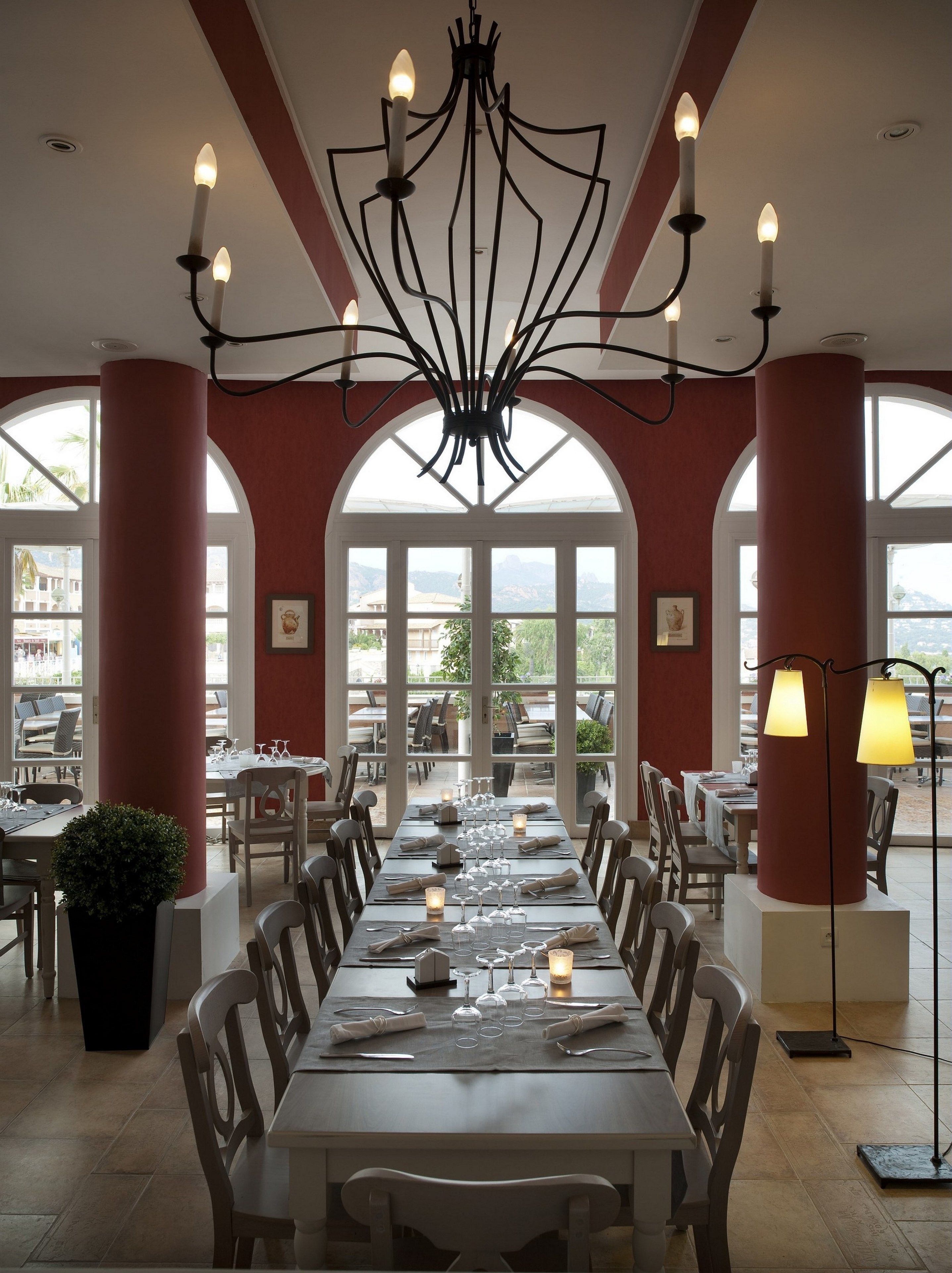 Un restaurant avec une longue table à manger dressée pour le repas, de grandes fenêtres et un lustre.