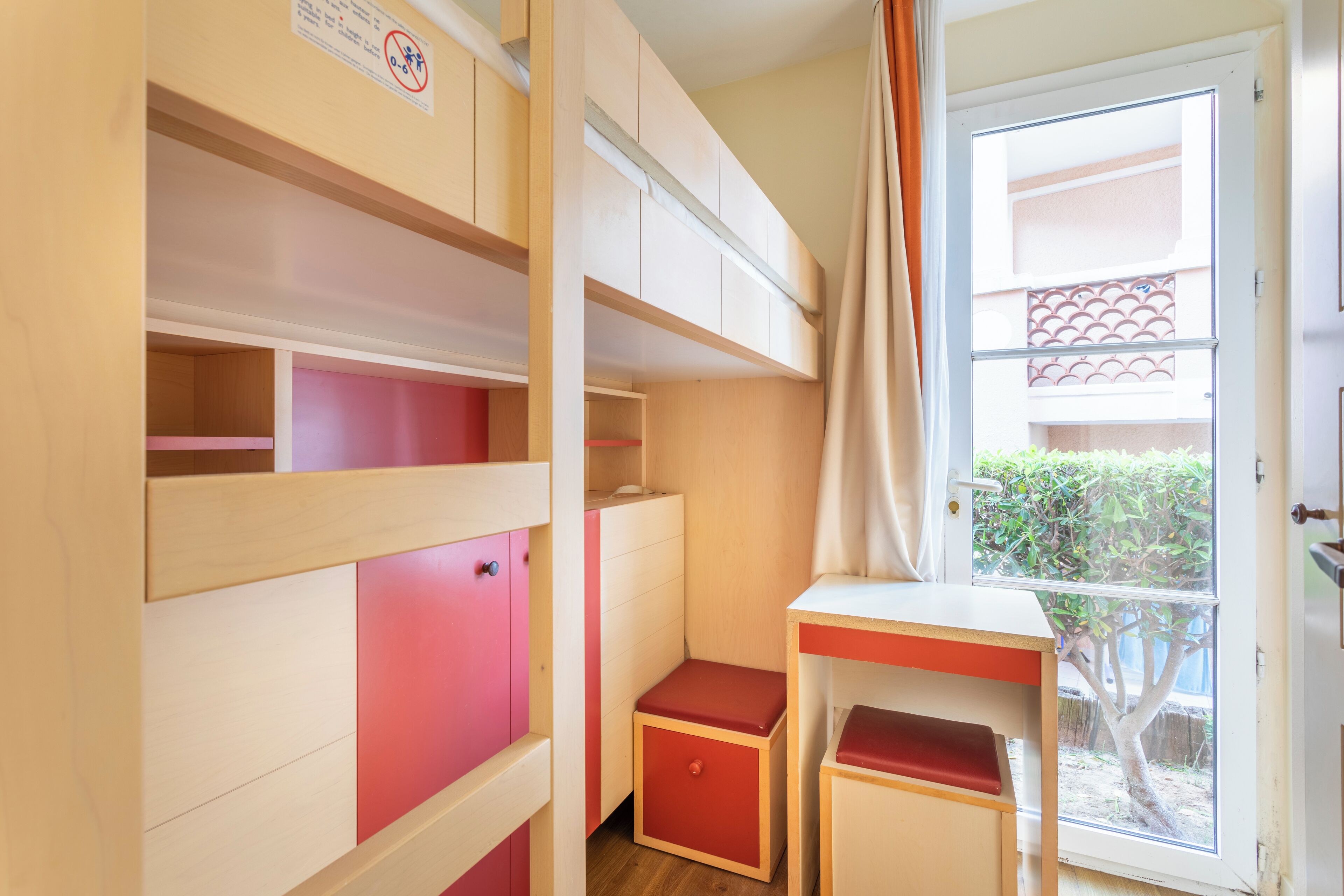 A compact room with bunk beds, a small desk, and a window with a view of a balcony.