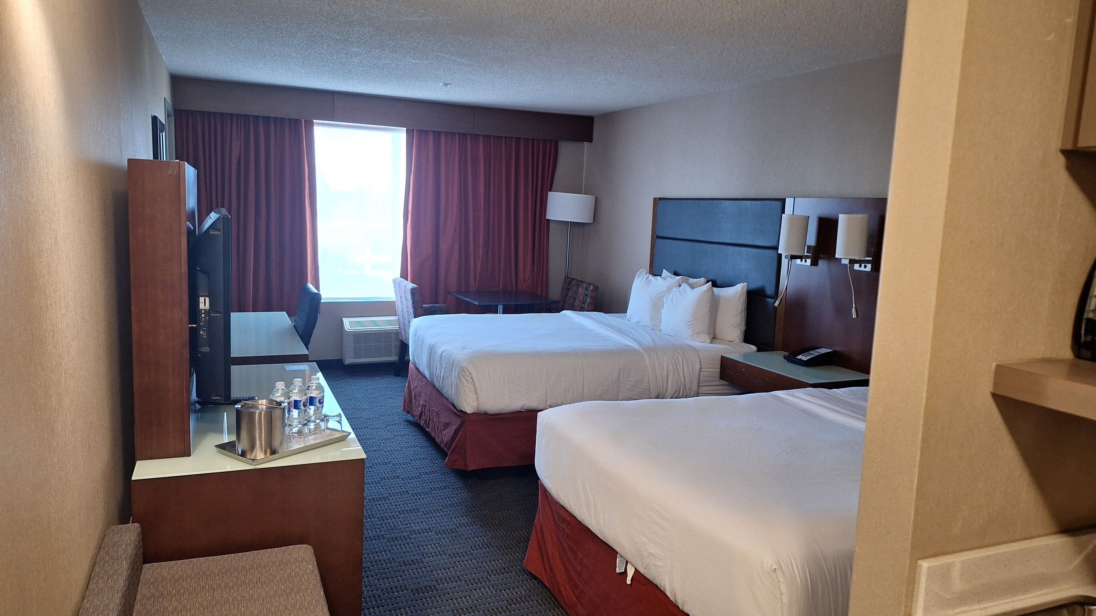 A hotel room with two beds, a desk with a chair, a TV, and a window with curtains.