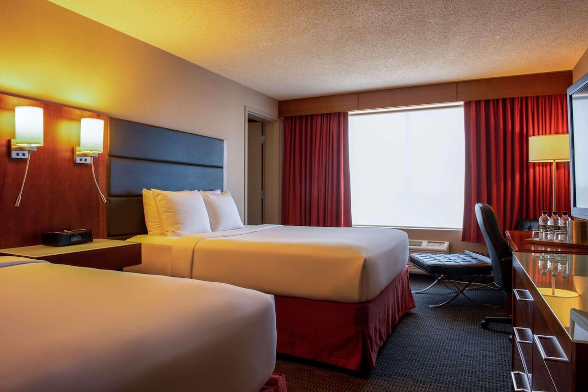 A hotel room with two beds, a desk, a chair, a desk lamp, and a large window with red curtains.