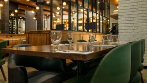 Un restaurante con mesas de madera y sillas verdes, ventanas amplias y lámparas colgantes.