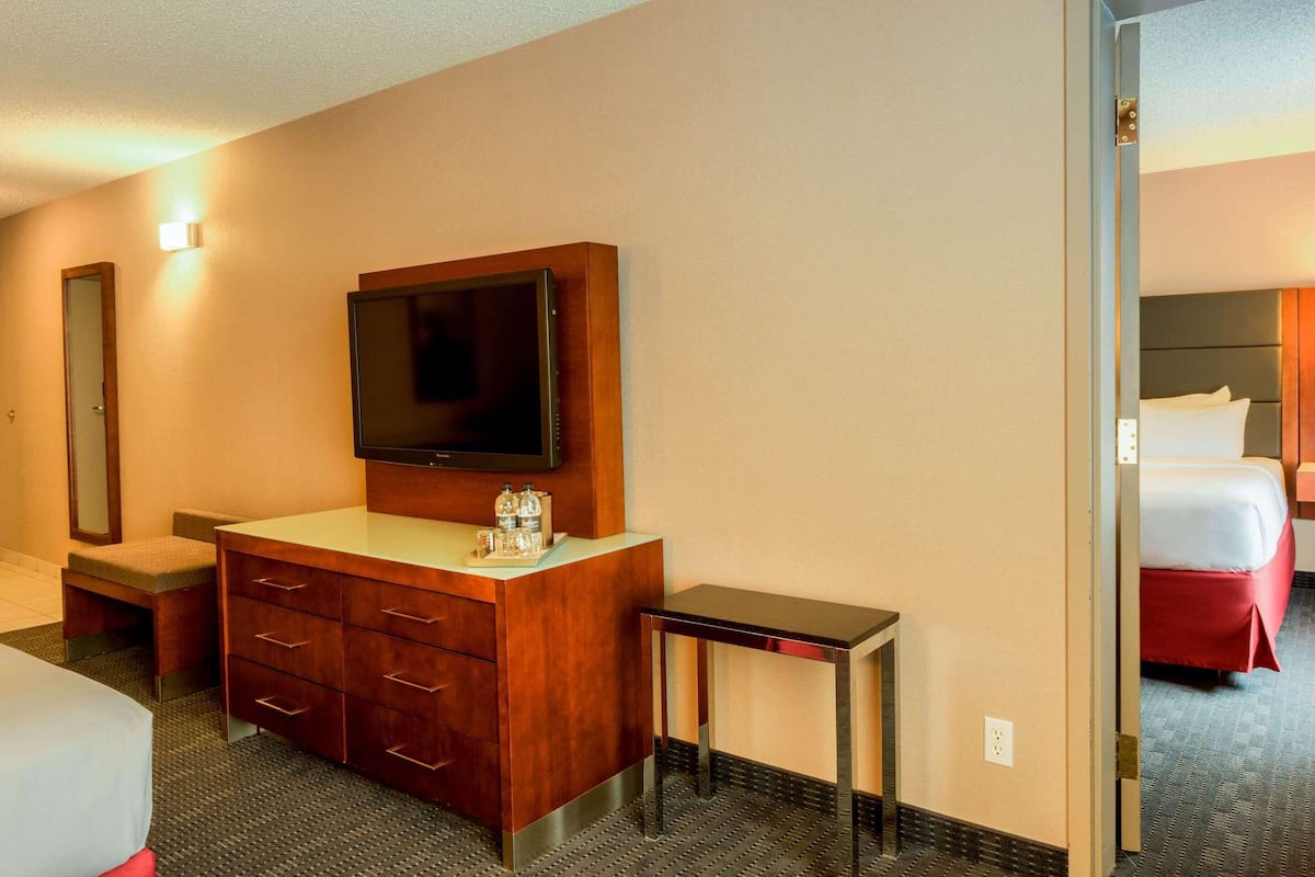 A hotel room with a wooden dresser, a flat-screen TV, a small table, a bed with red bedding, and a mirror.