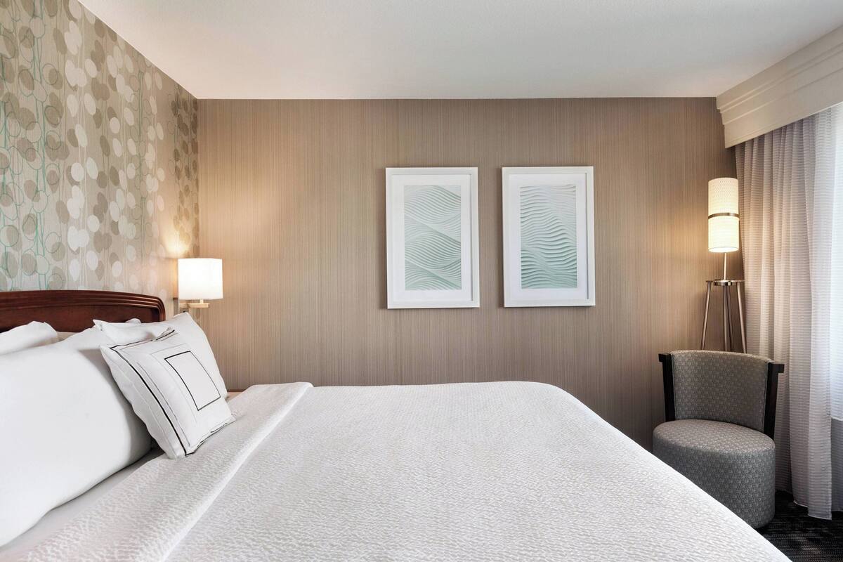 A hotel room with a large bed, two framed artworks on the wall, a chair, and a lamp.