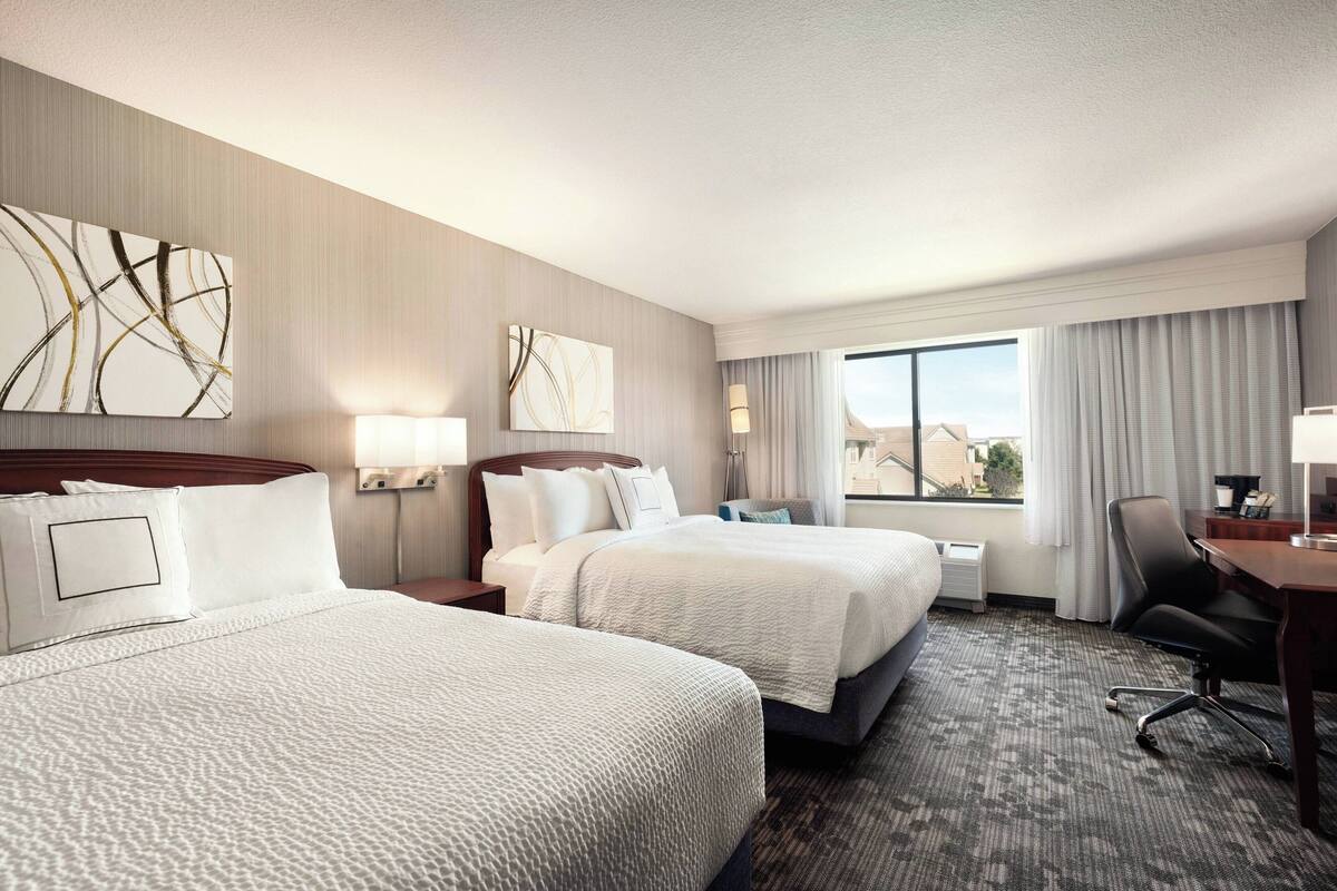 A hotel room with two beds, a desk, a chair, and a large window with curtains.