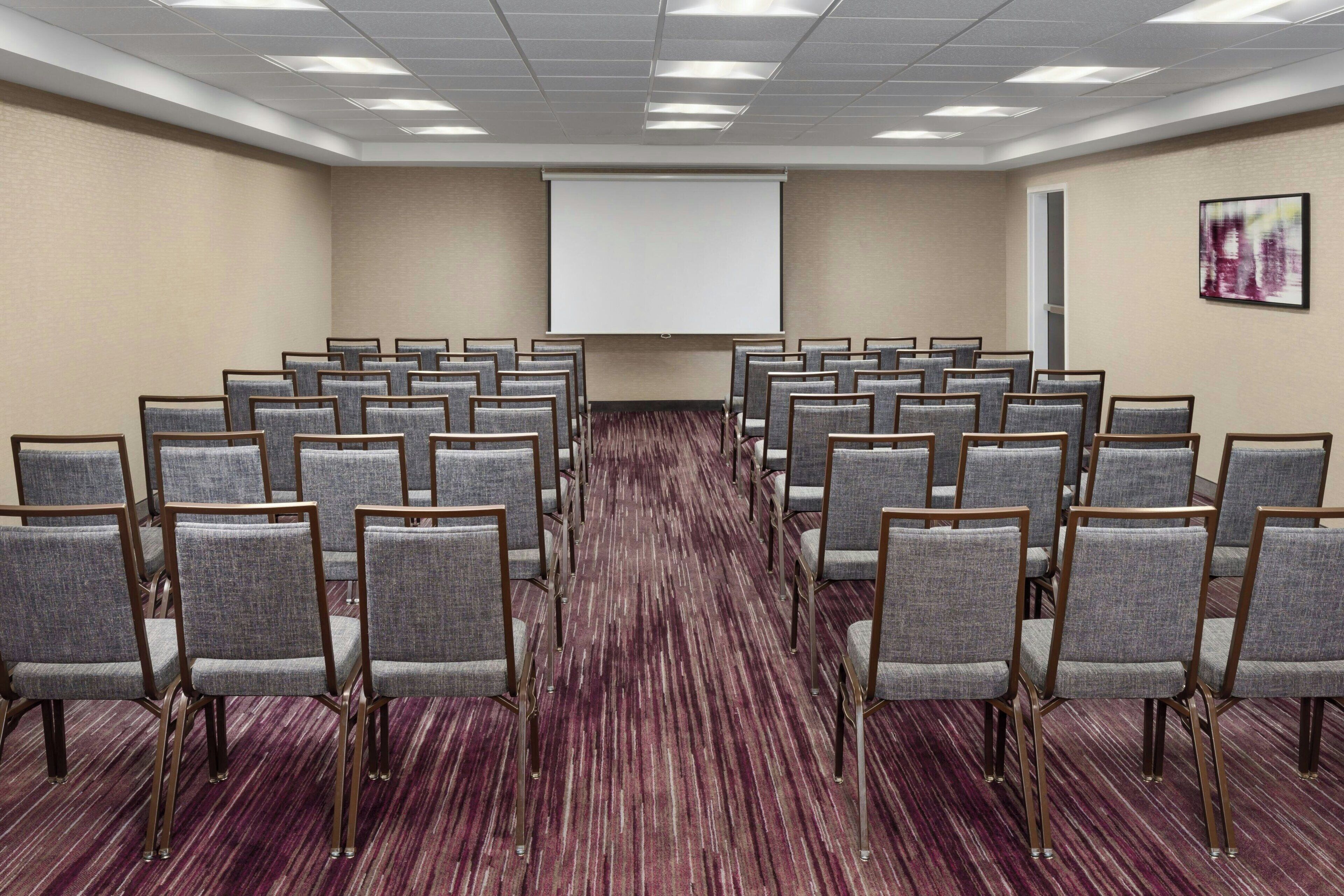 Une salle de conférence avec des rangées de chaises grises, un sol recouvert de moquette, un écran de projection et une porte sur le côté droit.