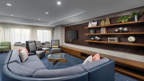 A modern living room with a sectional sofa, armchairs, a coffee table, and a TV mounted on the wall. There are shelves with decorative items and a large window with curtains.