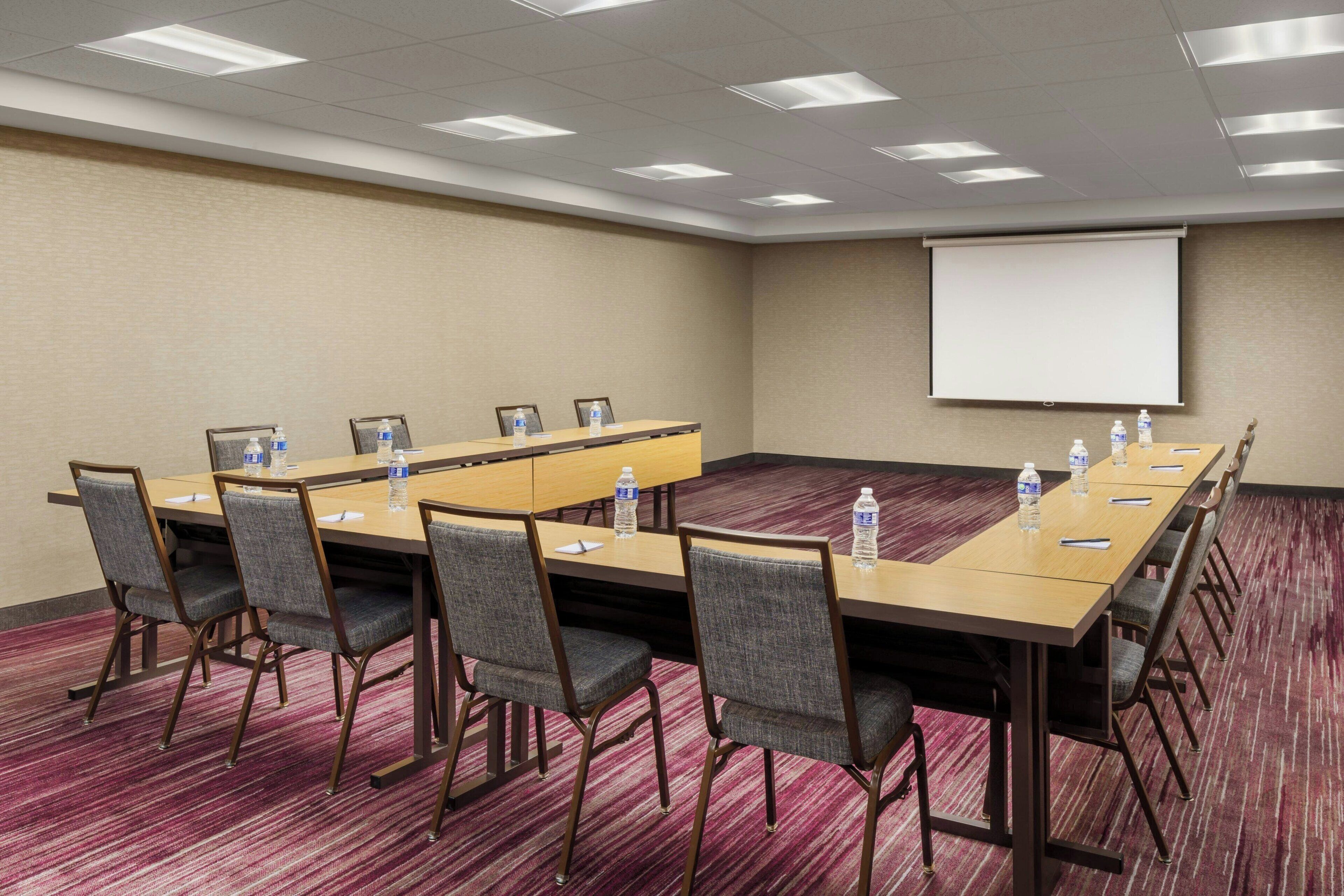 A conference room with a long table, chairs, a projector screen, and water bottles on the table.