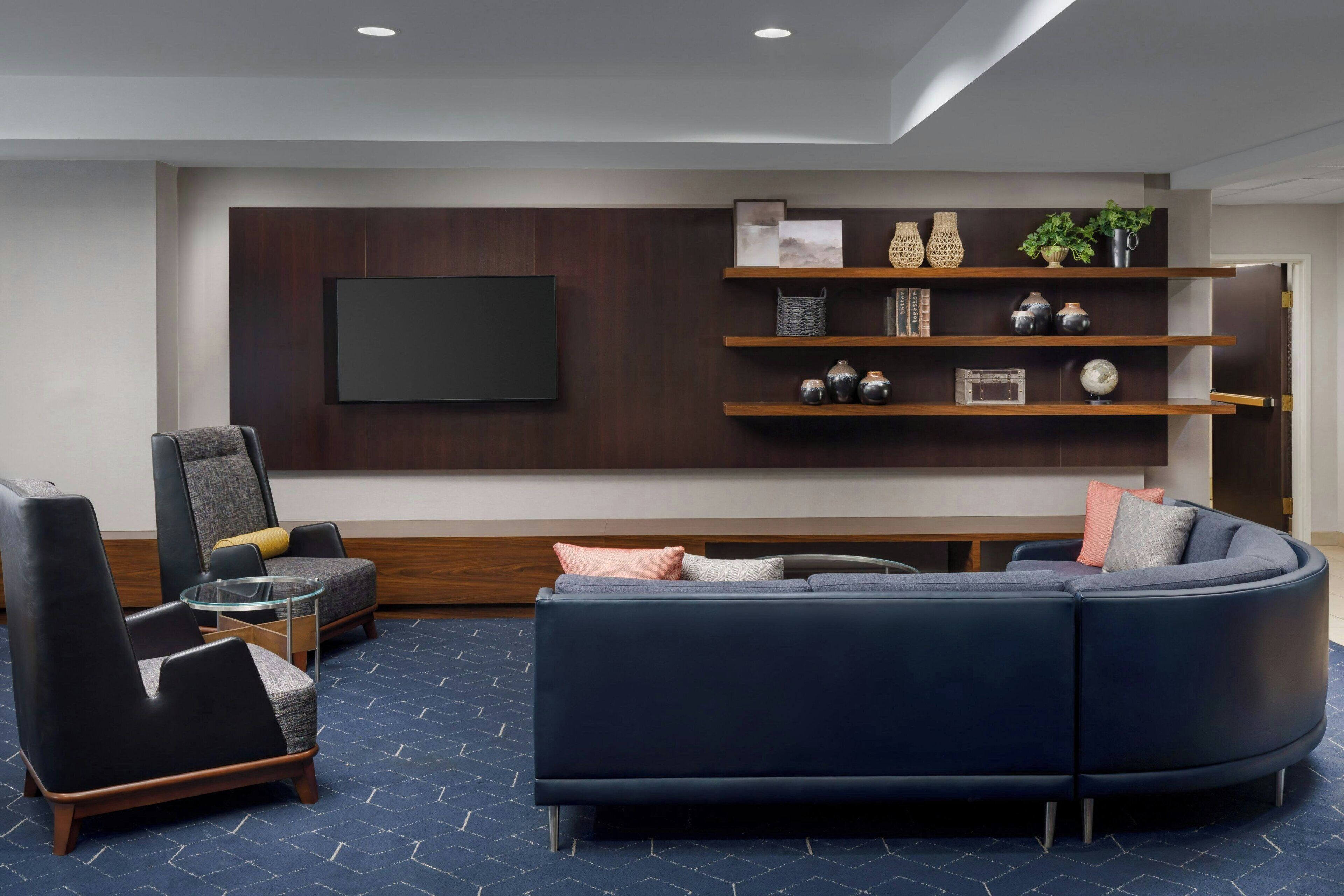 A modern living room with a dark blue sofa, two armchairs, a glass coffee table, and a flat-screen TV mounted on a wooden wall.