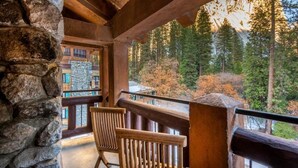 A balcony with stone walls and wooden chairs, overlooking a forested area with a mountain in the background.