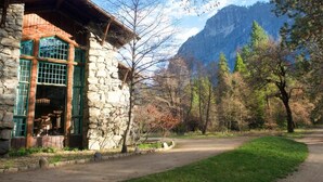 A stone building with a wooden roof and green-framed windows, surrounded by trees and a mountain in the background.