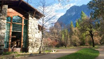 Un edificio in pietra con tetto in legno e finestre con cornice verde, circondato da alberi e una montagna sullo sfondo.