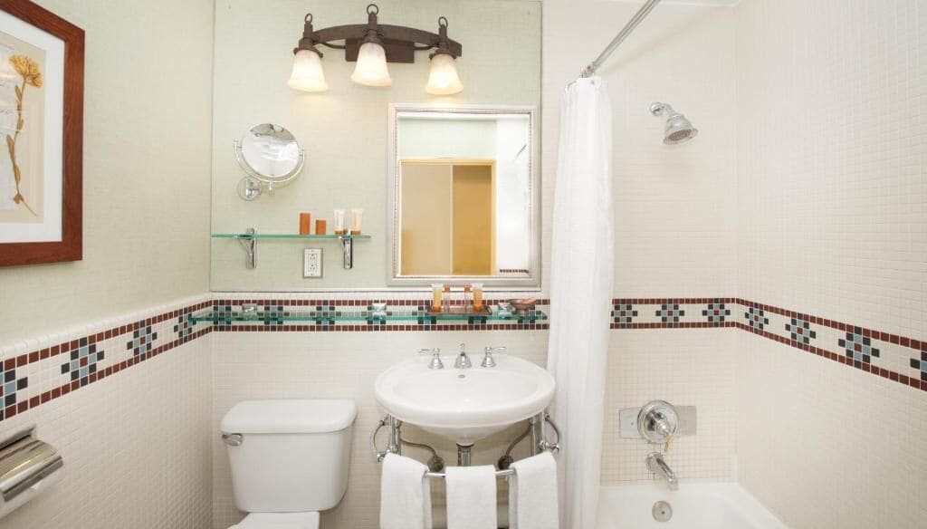A bathroom with a toilet, sink, and shower. There is a mirror above the sink and a framed picture on the wall.