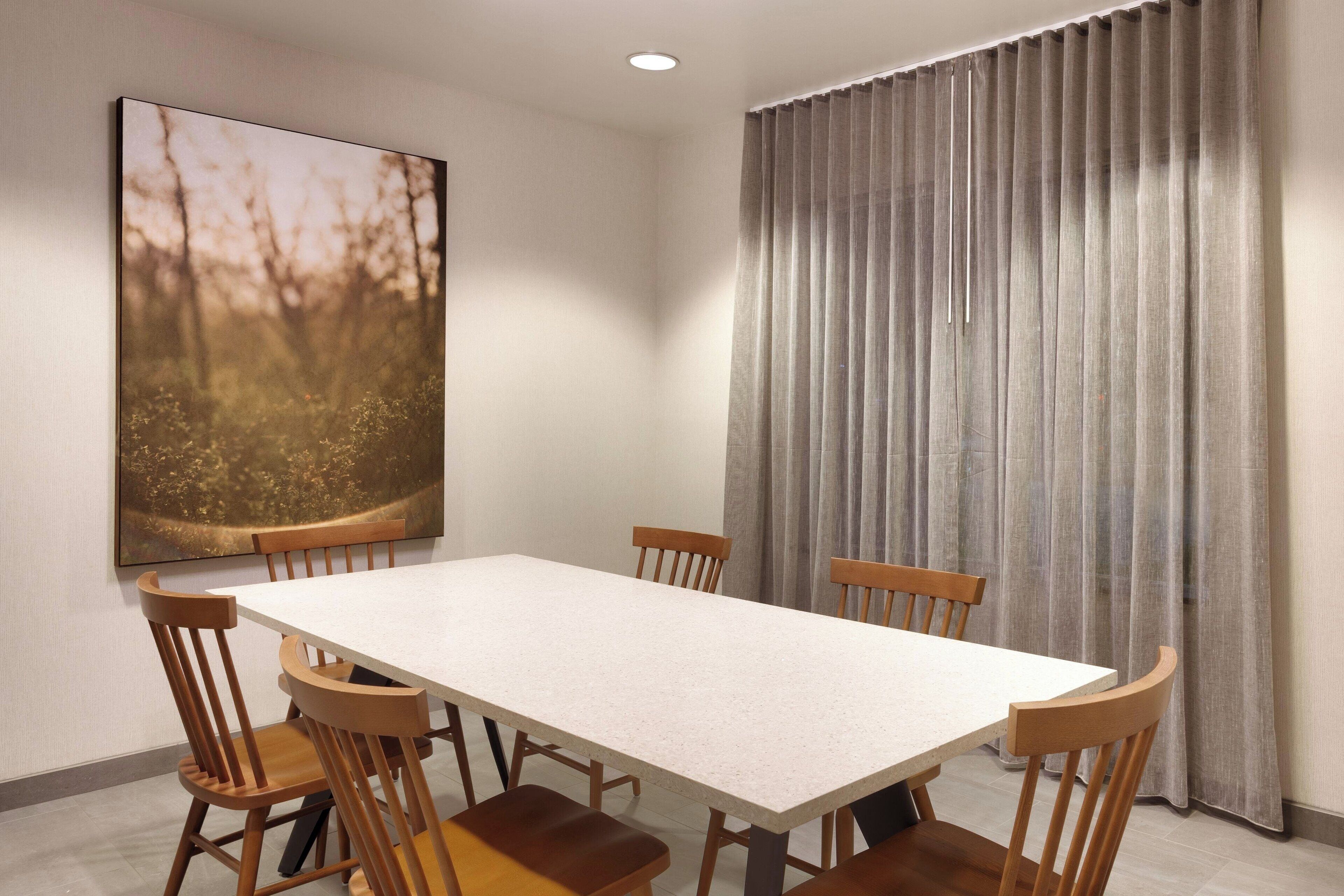 Comedor con mesa rectangular y seis sillas de madera. Un cuadro grande de un bosque adorna la pared, y una cortina gris cubre la ventana.