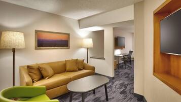 A hotel room with a sofa, a small table, a green chair, a painting, a lamp, and a TV.