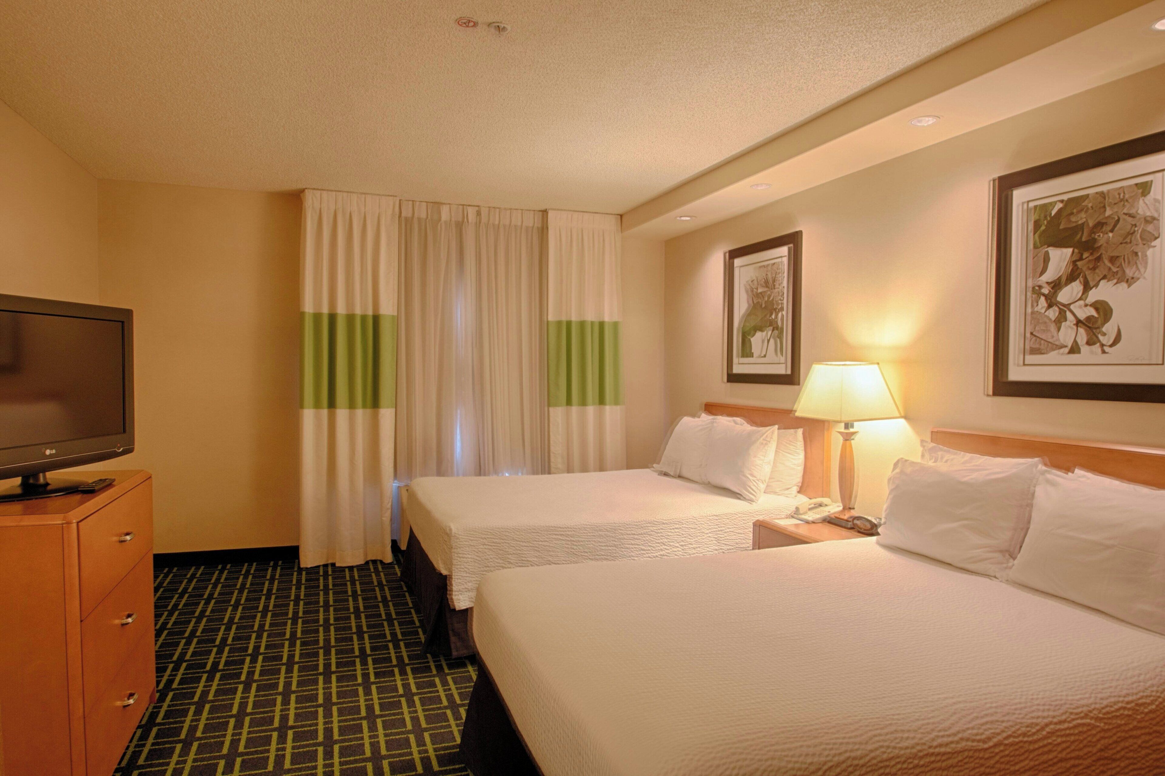A hotel room with two beds, a television, a desk with a lamp, and a framed picture on the wall.