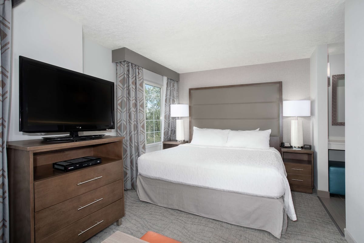 Una habitación de hotel con una cama grande, un televisor sobre una cómoda de madera y una mesita de noche con una lámpara.