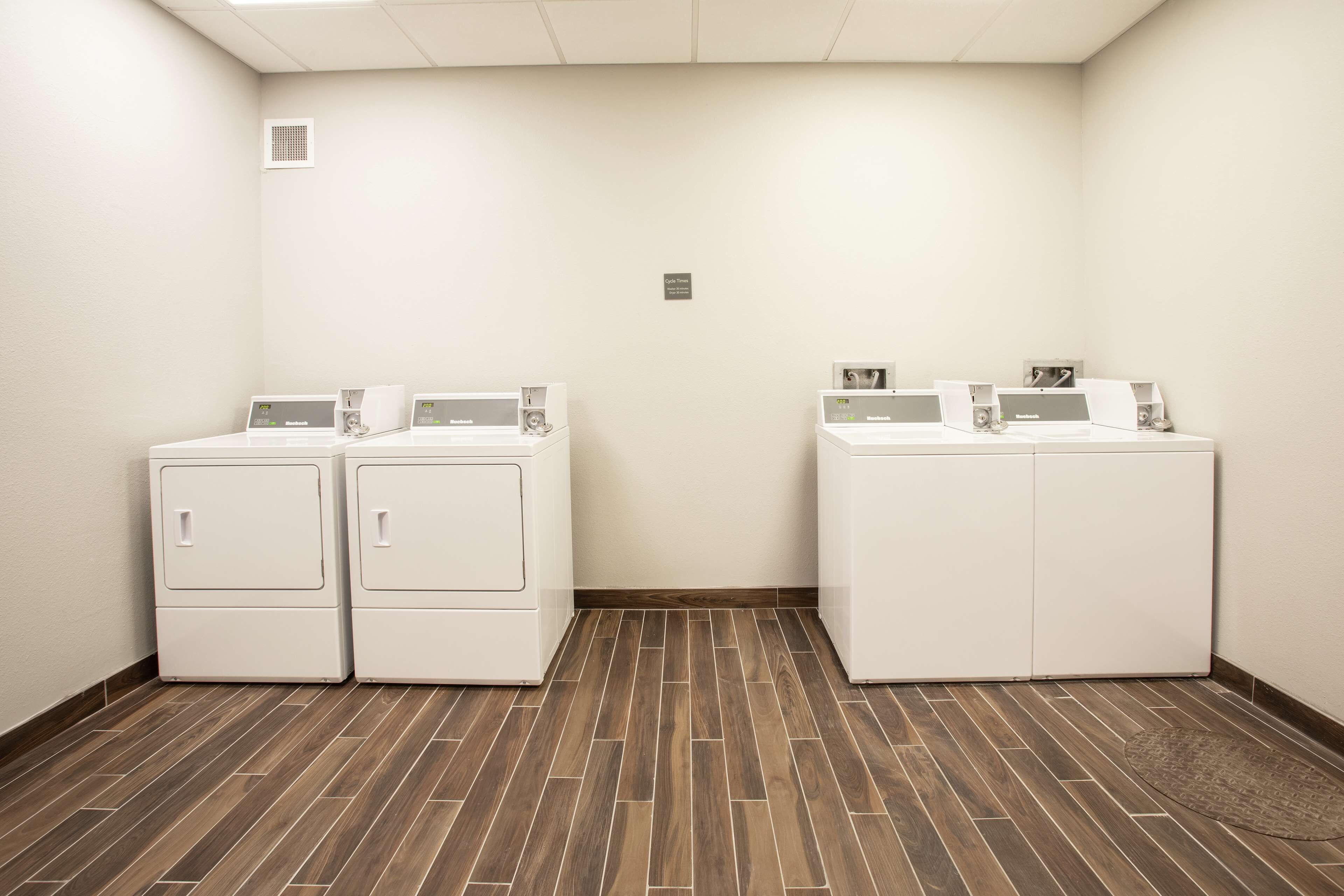 Een wasserij met twee rijen witte voorlaadwasmachines en drogers, een houten vloer en een kleine ventilatieopening in het plafond.