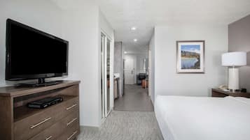 Ein Hotelzimmer mit Flachbildfernseher auf einer Holzkommode, einem Bett mit weißer Bettwäsche, einem gerahmten Bild an der Wand und einer Lampe auf einem Nachttisch.
