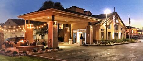 A hotel with a covered outdoor seating area, a well-lit entrance, and a landscaped front yard.