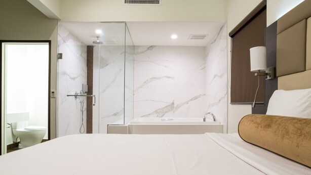 A modern hotel room with a large bed, a glass-enclosed shower, a marble wall, and a bedside table with a lamp. - Hotel Xilo Glendale (Glendale)