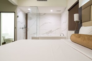 A modern hotel room with a large bed, a glass-enclosed shower, a marble wall, and a bedside table with a lamp. - Hotel Xilo Glendale (Glendale)